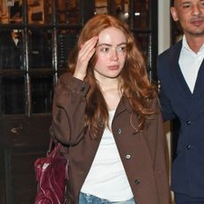 Sadie Sink wearing a Prada brown jacket, white tank top, blue bootcut jeans, burgundy bag, and black boots