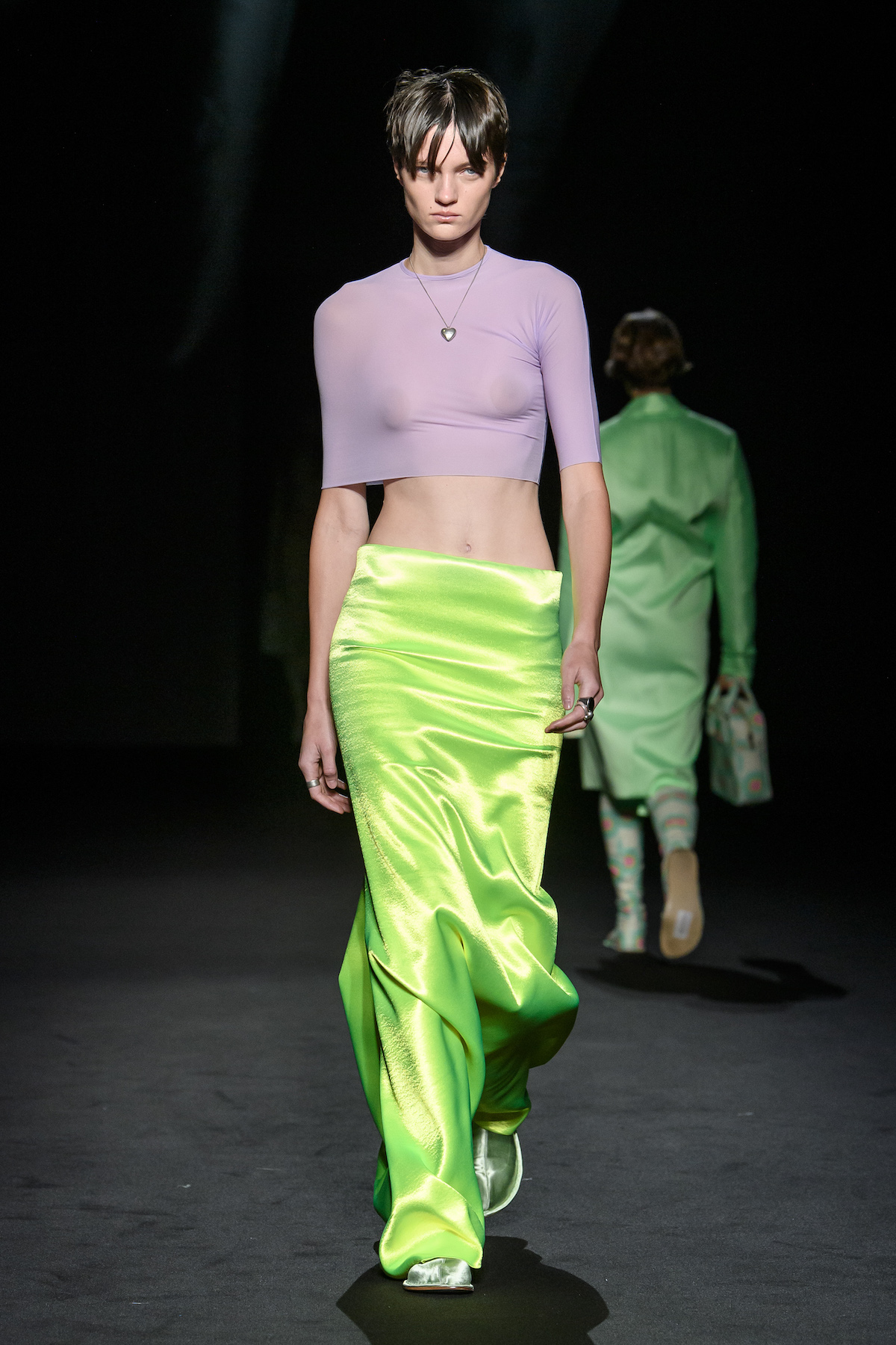A female model wearing a short purple top and long shiny green pants walking down a runway.