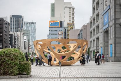 A street level view of a piece of street art in the middle of a street plaza. The art is a bowl shape structure with open sides that is made from wood and is c. 12ft high. 