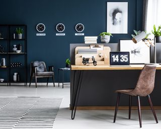 Dark blue home office with wooden desk, brown leather chair and three clocks on the wall in the background
