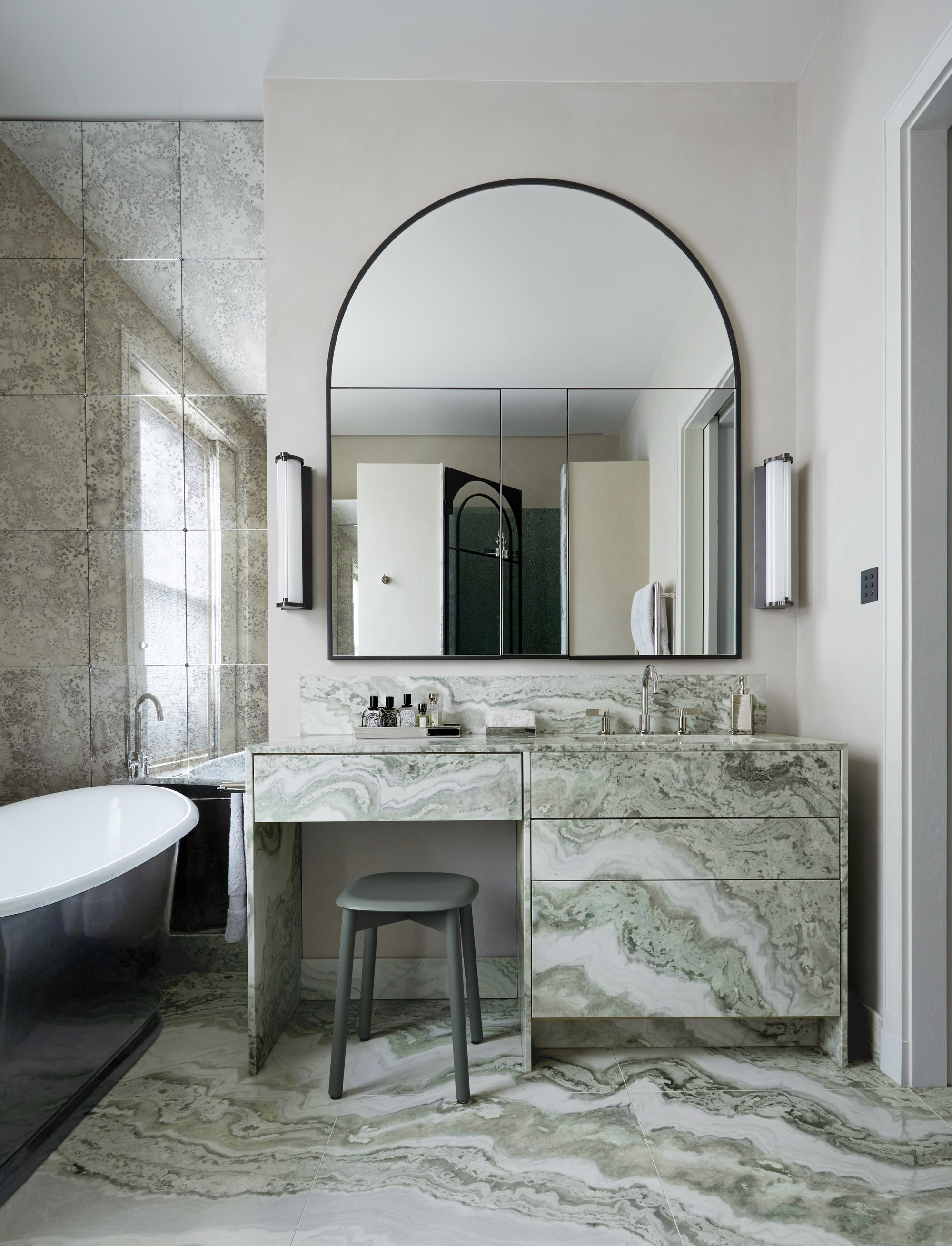 green marble bathroom with marble across vanity, drawers, and floor, with mirrored wall next to freestanding bath, and vanity stool in front of large arched mirror