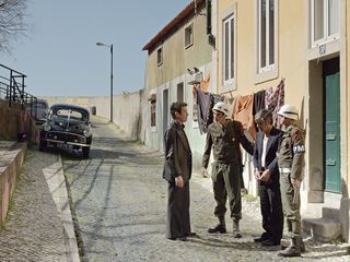 Image shows a still from the film 'The Secret Agent', a group of men talking in a street