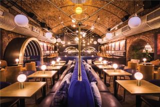 A night venue features warmly lit spherical lights placed on each of its square wooden tables, vevlet and leather soft seating, and vibrant brick walls in an imaginative revisiting of an industrial site.