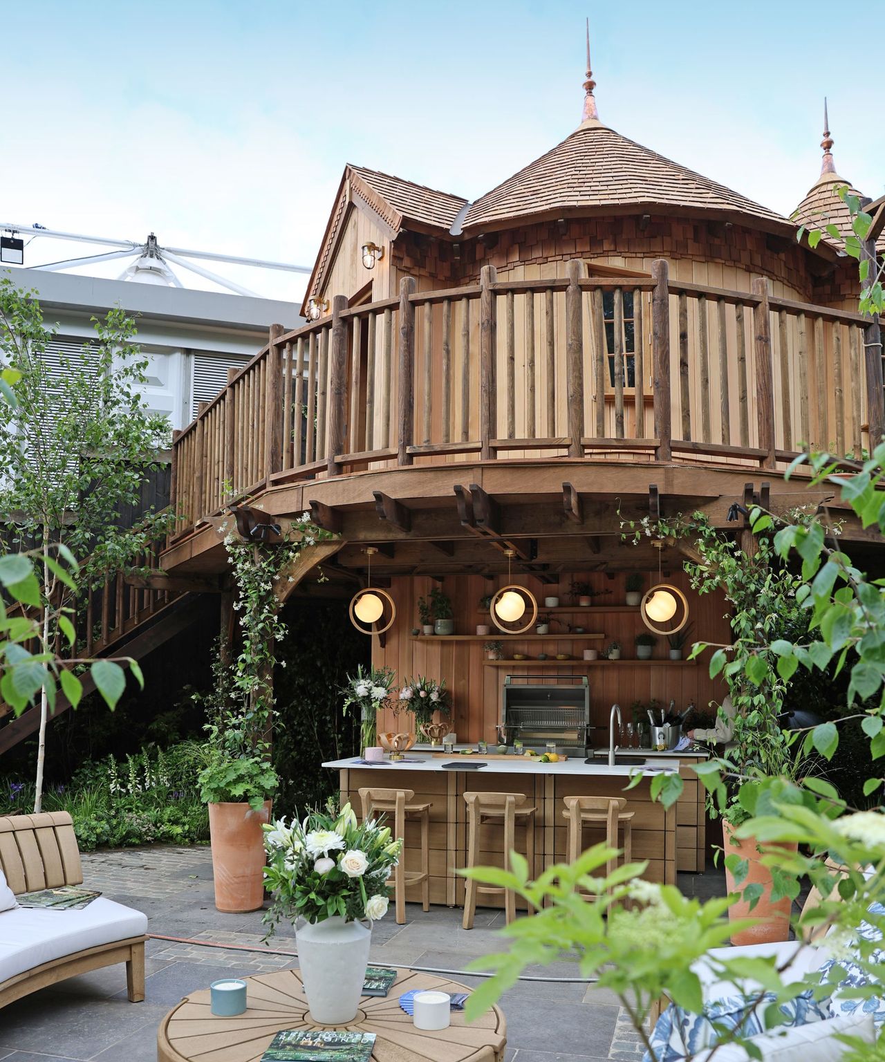 A beautifully crafted timber treehouse with a wraparound balcony, shingle roof and turret-style details. Below, a fully equipped outdoor kitchen and bar with pendant lighting sits among lush planting.