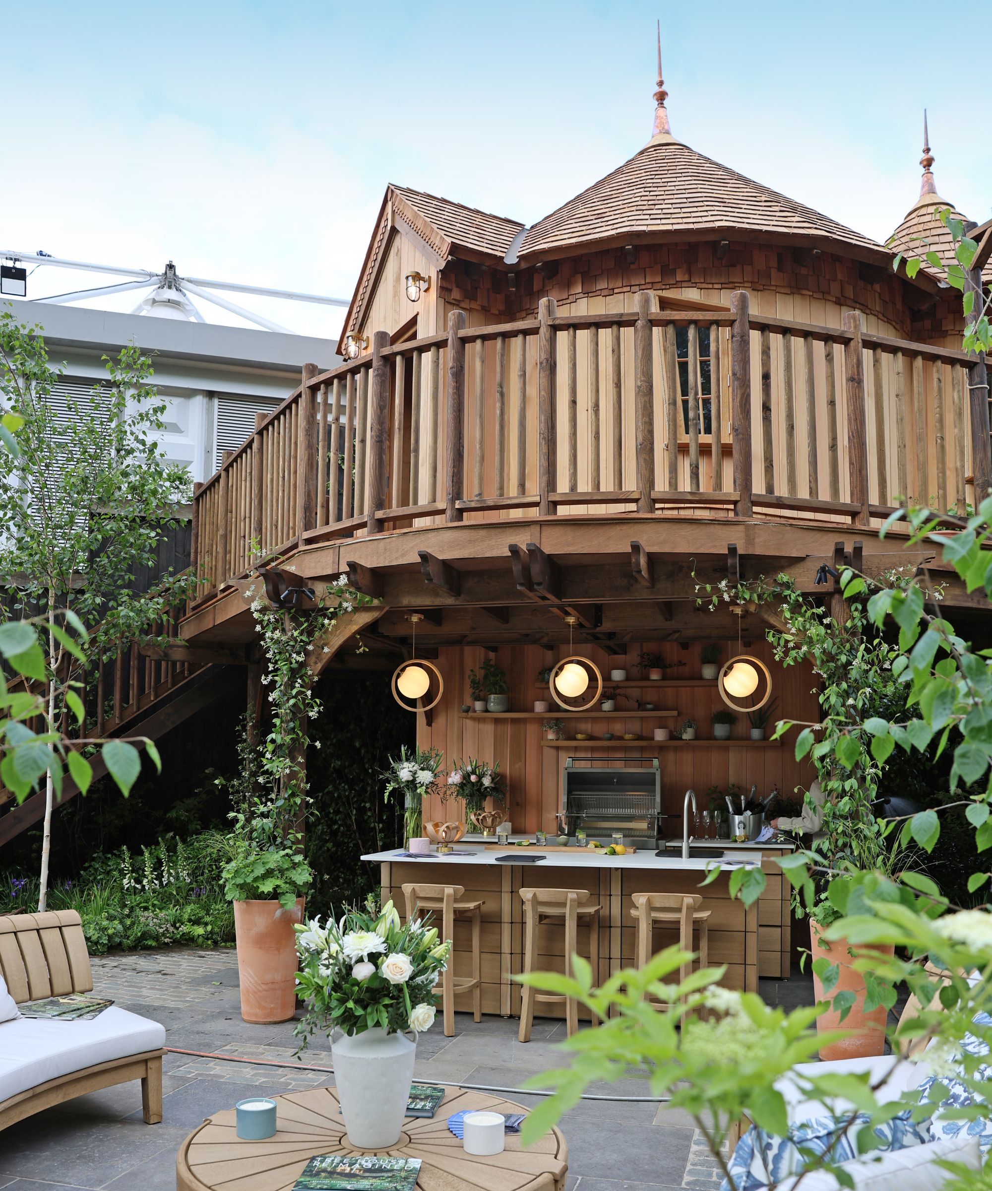 A beautifully crafted timber treehouse with a wraparound balcony, shingle roof and turret-style details. Below, a fully equipped outdoor kitchen and bar with pendant lighting sits among lush planting.
