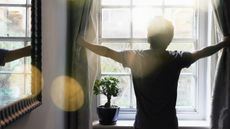 A man opens the curtains over a window where bright sunlight shines through.