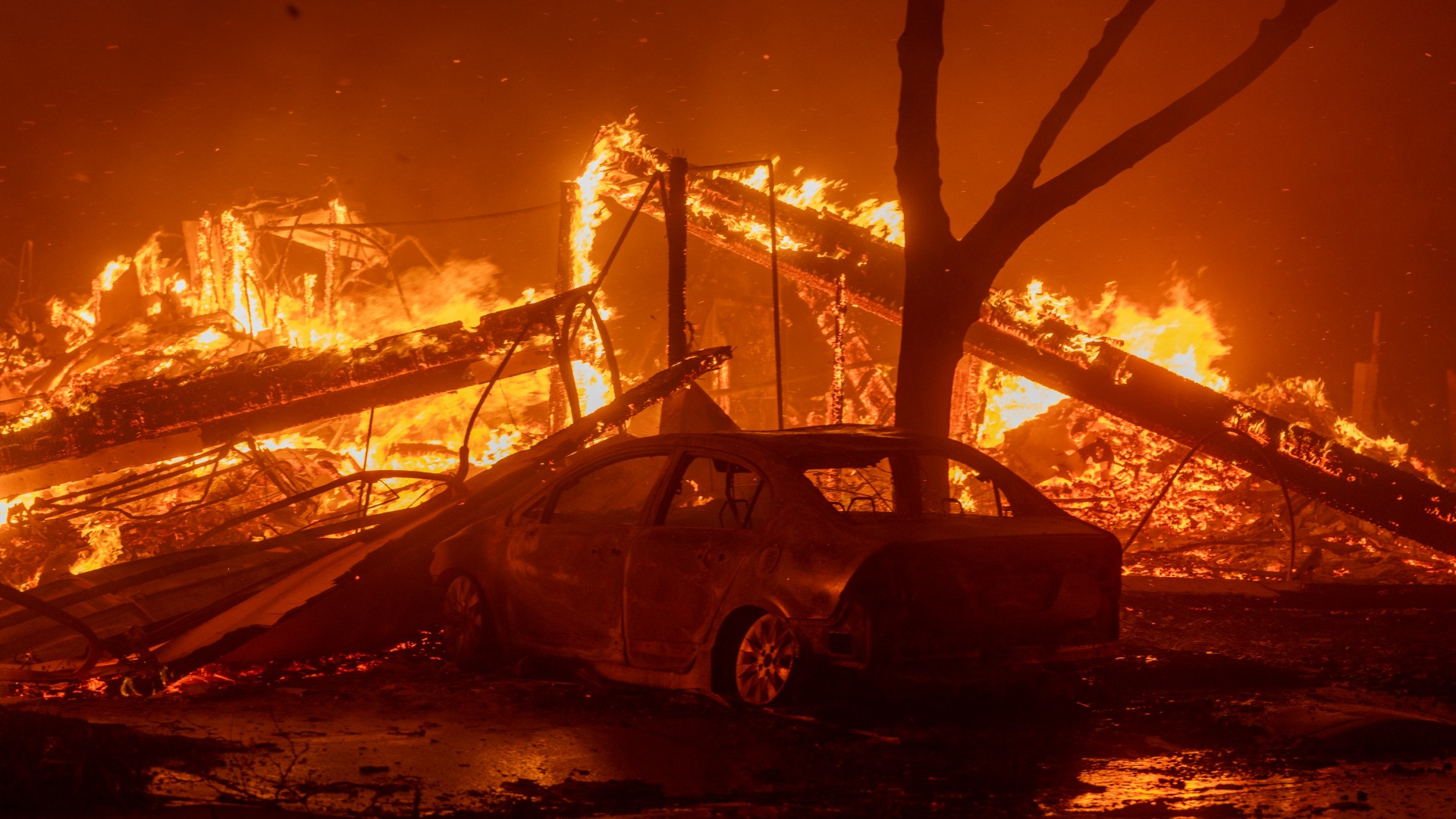 a collapsed house is engulfed in flames atop a melted car