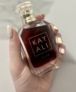 Hand with pale yellow nail polish holding up kayali vanilla 28 perfume in dark brown bottle with gold and clear jewel-shaped cap on grey and white marble background