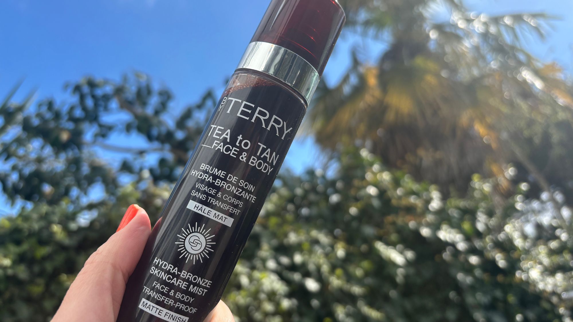 a hand holding a bottle of by terry tea to tan face and body in the sunshine