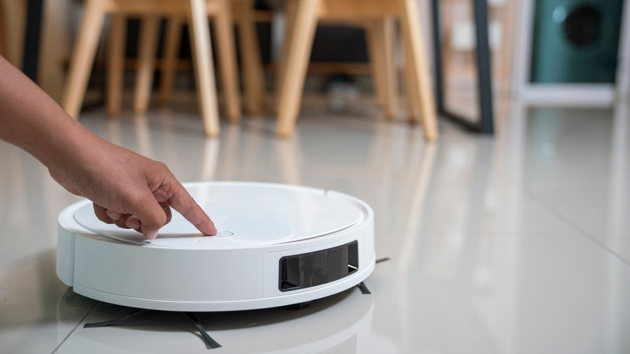 White robot vacuum on a glossy grey tiled floor with hand reaching in to press a button
