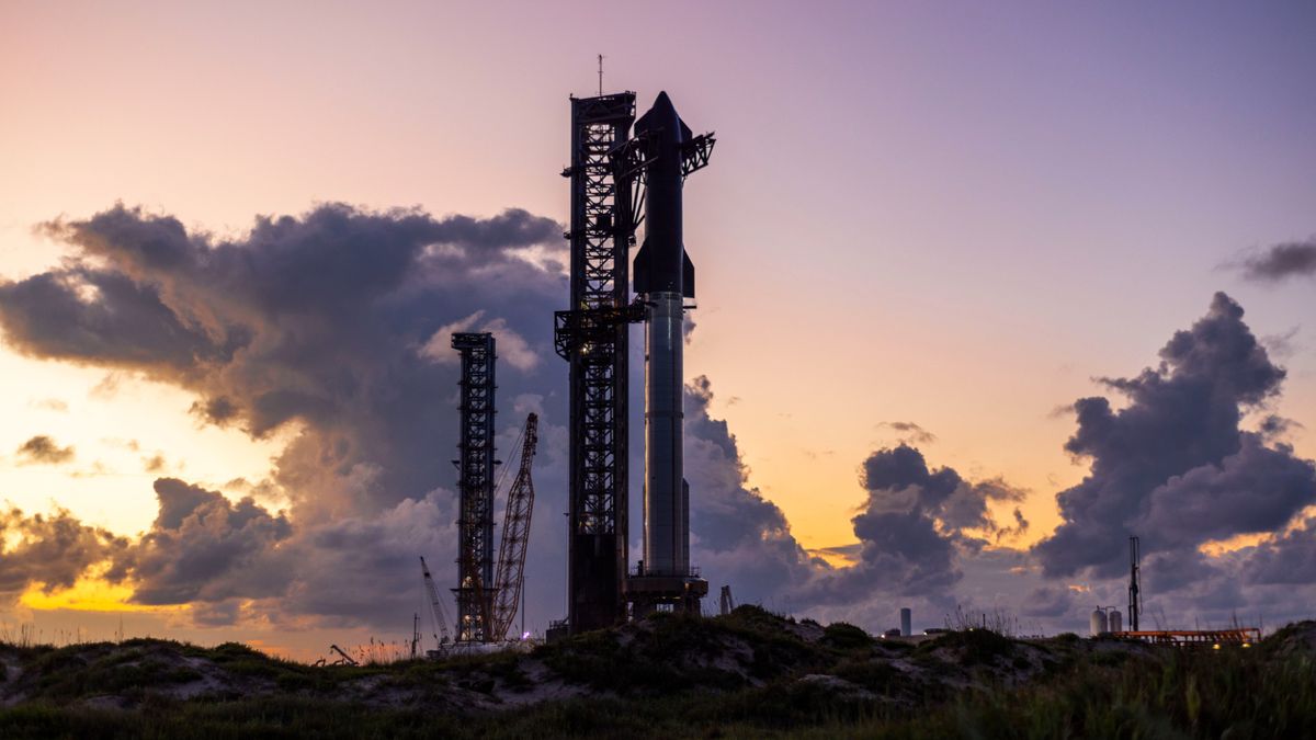 A massive rocket stands next to a launch tower before a brilliant sky of tall purple clouds.