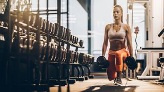 Woman performs lunge movement with dumbbells next to rack of dumbbells in a gym