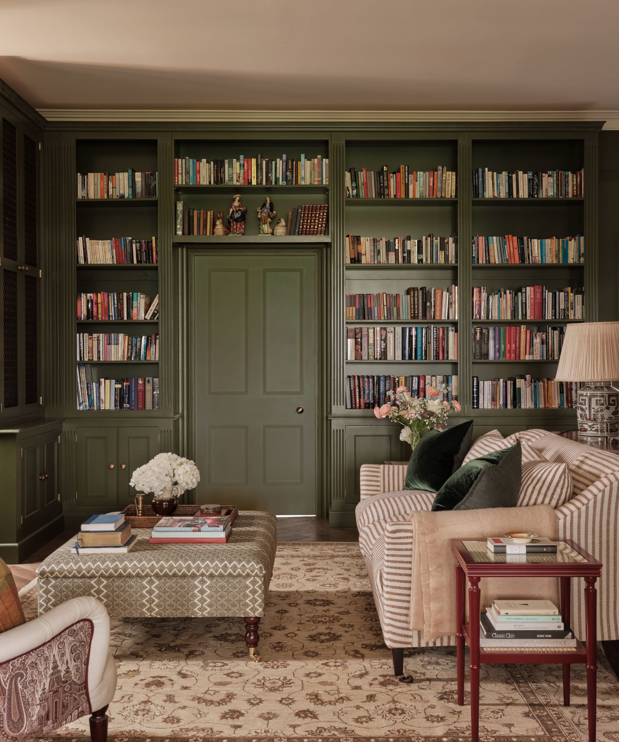 Living room with dark green painted built-in shelving holding bookcases, a striped beige and cream sofa, a patterned ottoman, a red side table and a neutral Persian rug