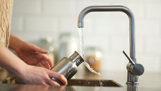Reusable tin water bottle under tap in kitchen with running water going into it, an example of alternatives to caffeine