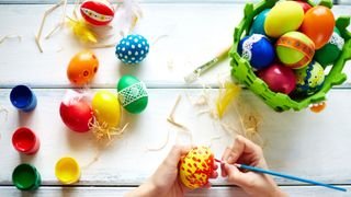 Image of colourful eggs strewn across a table and a childs hand painting them as a suggested Easter game