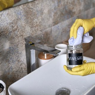 Bottle of vinegar being used to clean around bathroom sink, grey wall tiles with white grouting