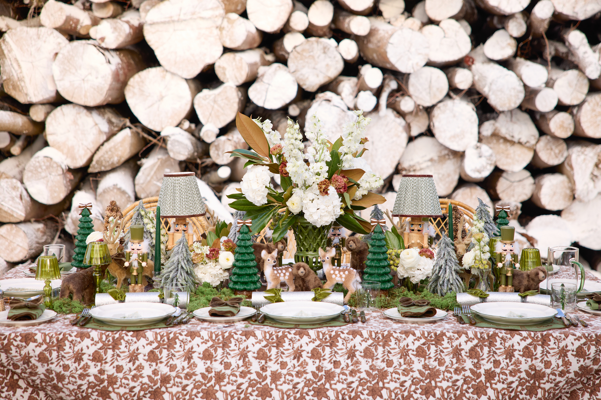 whimsical christmas table setting with fawns, patterned tablecloth, stack of wood behind, and green trees, with table lamps