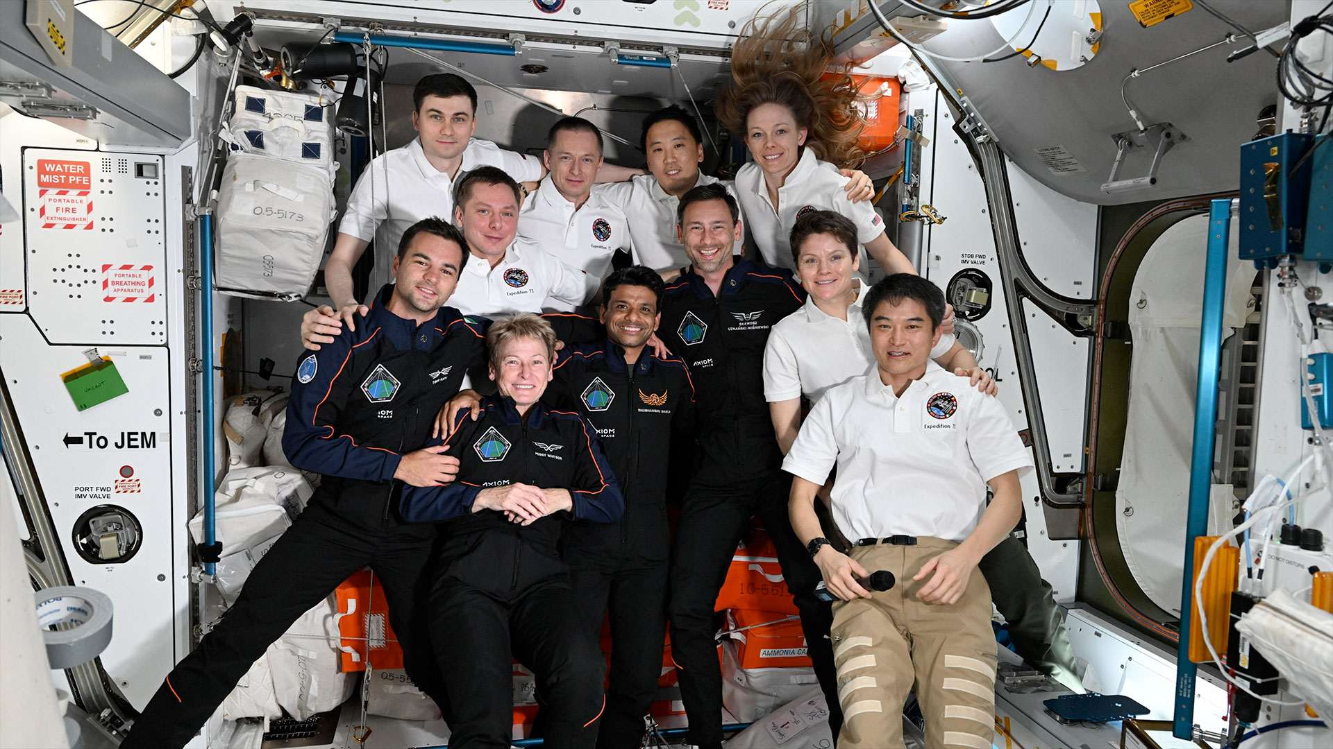 Eleven people, including three women, gather together aboard a space station for a group photo. Seven are wearing white polo shirts and kakhi pants. Four are wearing dark blue flight suits.