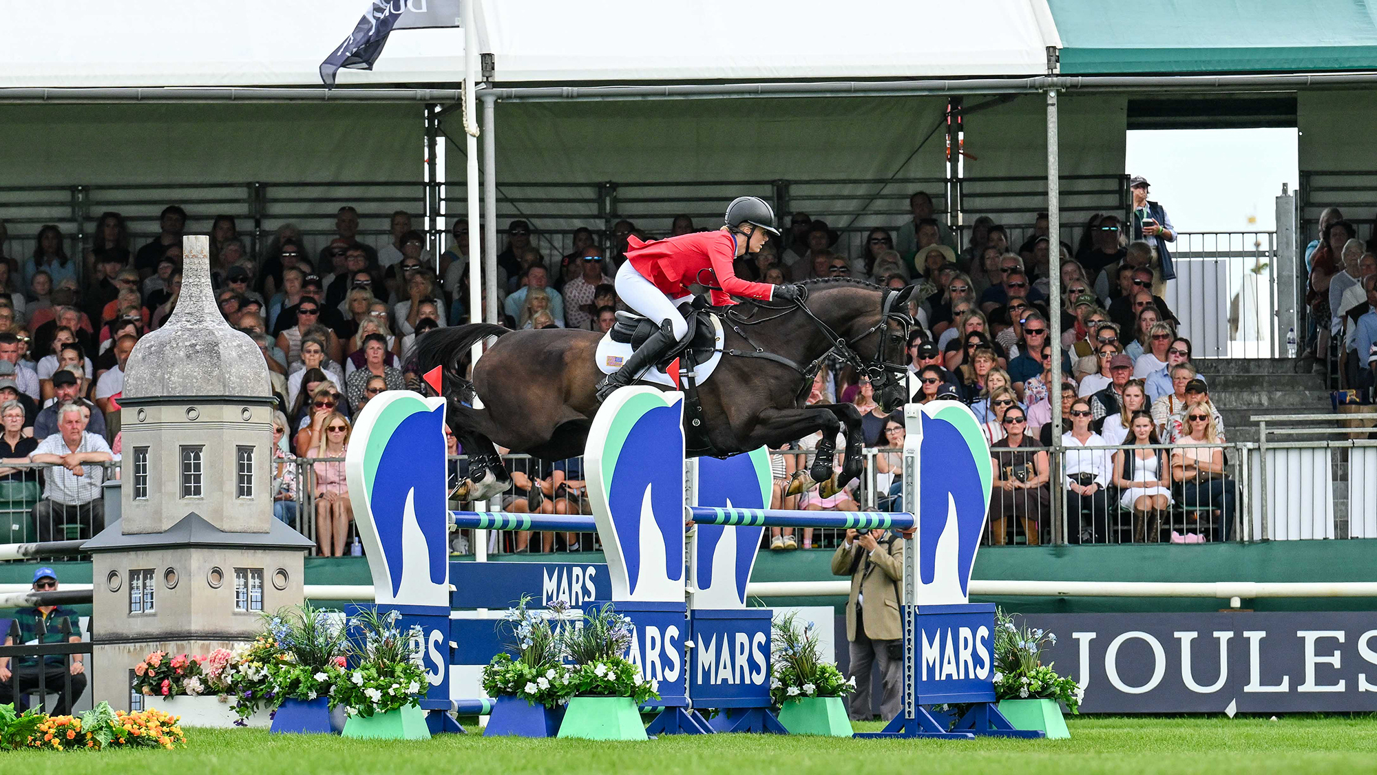 Caroline Pamukcu, a rider in a red coat, rides black horse HSH Blake over a blue and green showjump at Burghley