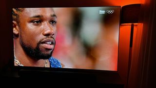 Close up of USA sprinter Noah Lyles from the Paris 2024 Olympic games, shown on the LG G4 TV. A floor lamp is to the right, emitting the colour red.