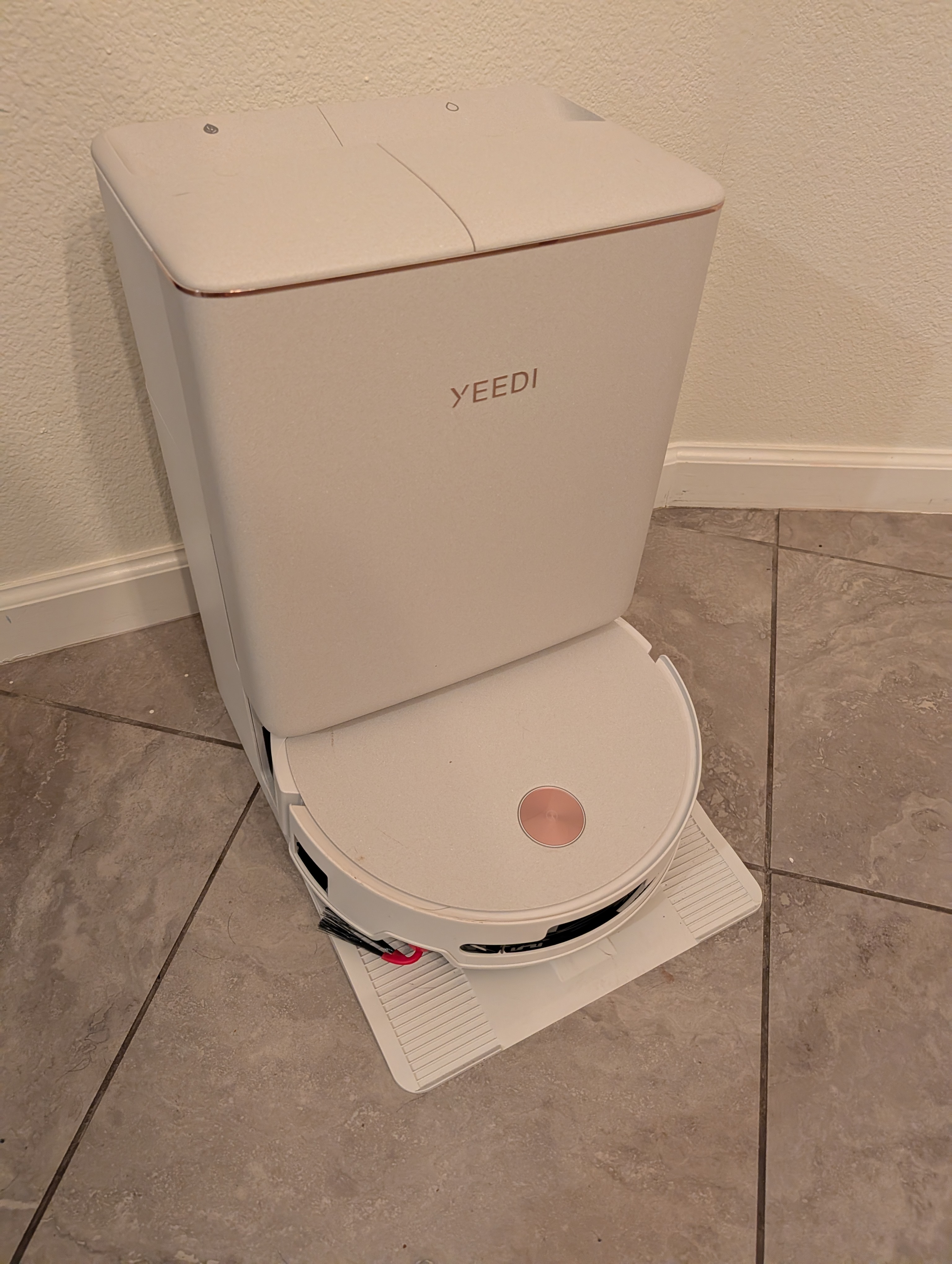 The Yeedi M16 Infinity docked in its station on a tile floor against a white wall.