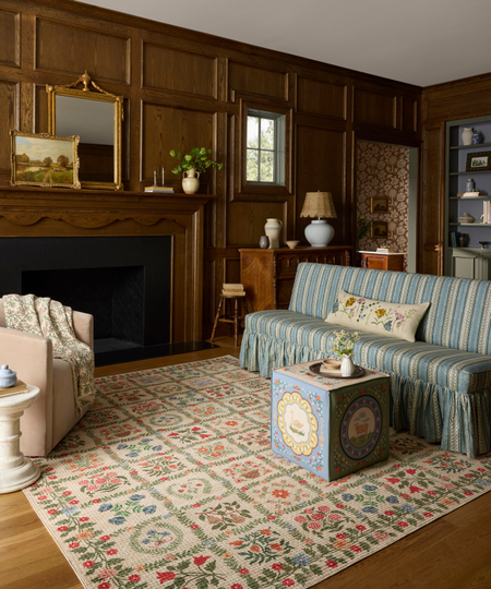 a cozy living room with wood paneled walls, a printed rug, a couch with stripes, a square ottoman, and a large fireplace