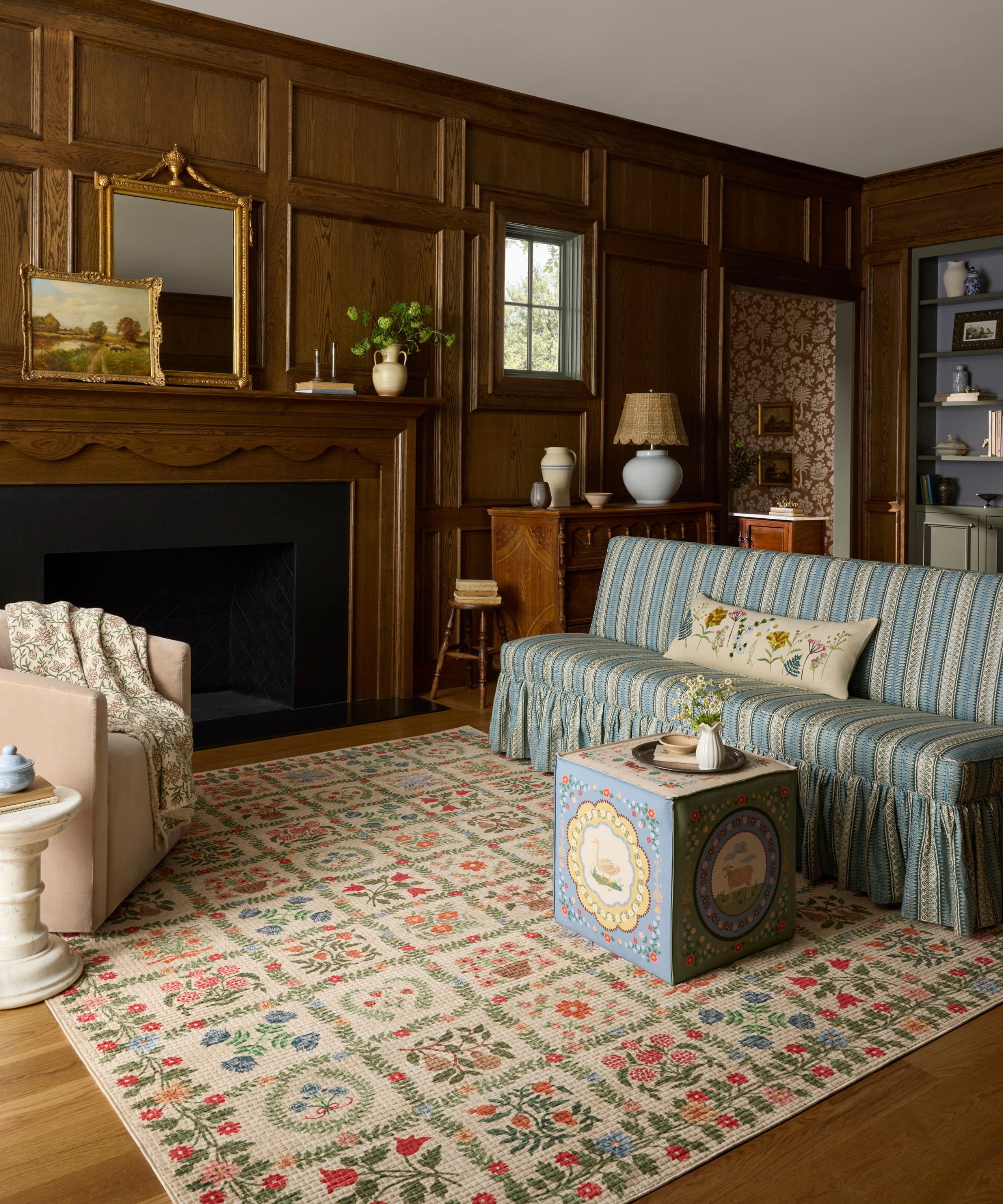 a cozy living room with wood paneled walls, a printed rug, a couch with stripes, a square ottoman, and a large fireplace