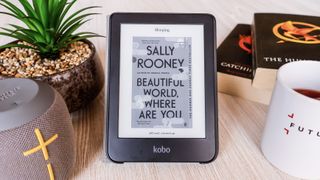 The Kobo Clara 2E positioned between a speaker, fake plant, two books and a coffee mug. On the ereaders display is an image of the cover of the current book being read on the device.