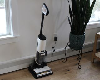 Tineco Floor One S7 steam wet-dry vacuum cleaner plugged into mains power socket on laminate floor with indoor plant to right