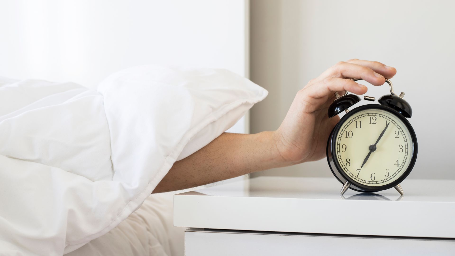 A person reaches their hand out to their alarm clock from under their comforter.
