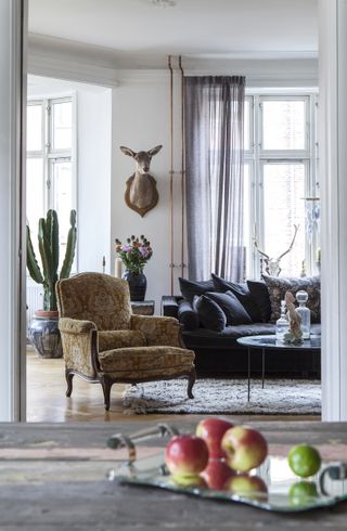 A Danish sitting room with decorative armchair, stag taxidermy wall decor, dark upholstered sofa, cream rug and vase with bouquet of flowers