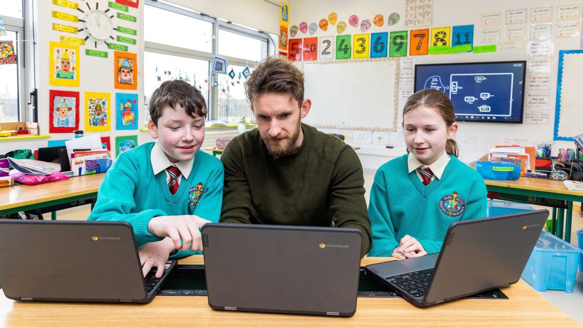 Two young students and a teacher huddle around computers in a classroom.