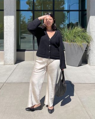 Female curvy fashion influencer poses on a sunny sidewalk shielding eyes from the sun wearing a black v-neck cardigan sweater, woven shoulder bag, cream satin pants, and black ballet flats.
