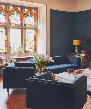 Living room with dark blue walls, wooden floor and dark blue velvet armchairs and sofa, leaded stone window with stained glass in the background.