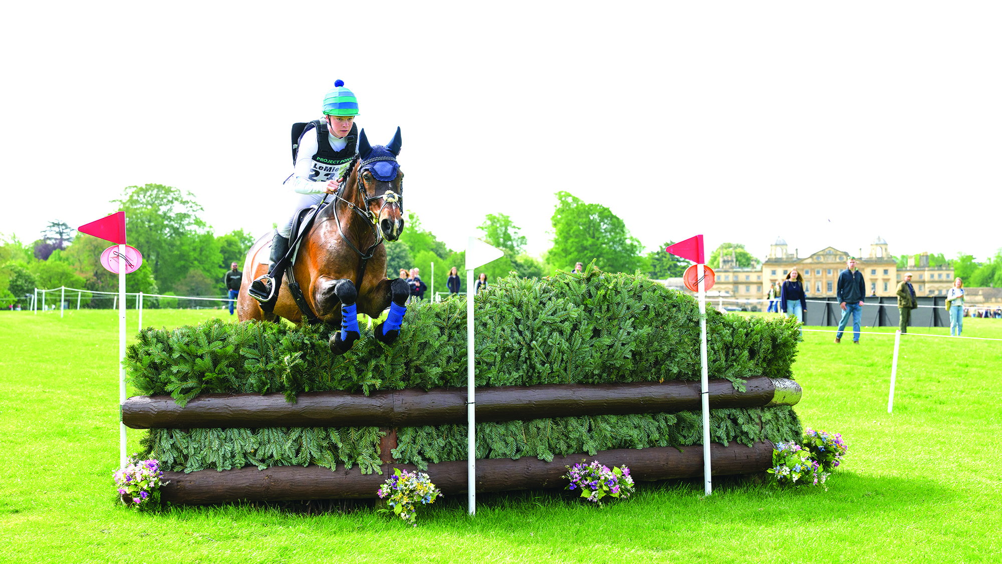A bay horse clears a large BE100 brush fence on the LeMieux Grassroots course with Badminton House in the background.