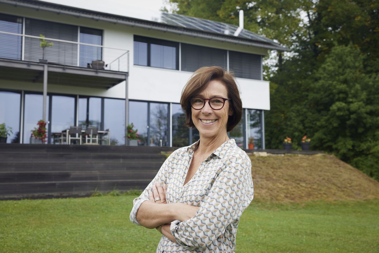 A 60-year-old woman in glasses stands similing in front of her house, folding her arms.