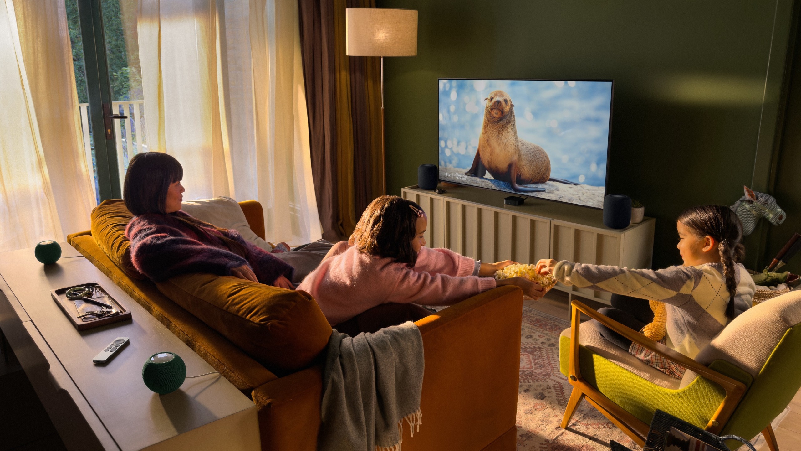 A family watching a nature documentary on TV in a modern living room, with HomePod speakers flanking a TV and two HomePod Mini speakers behind them.