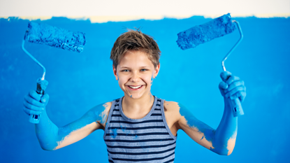 Portrait of happy teenage boy holding two paint rollers with his arms covered in blue paint.