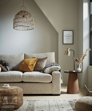 neutral living room with grey couch with cushions and throw, with a wicker coffee table and ceiling lampshade as well as a small side table with gold and black lamp