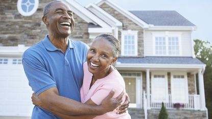 An older couple happily embrace in front of their house.