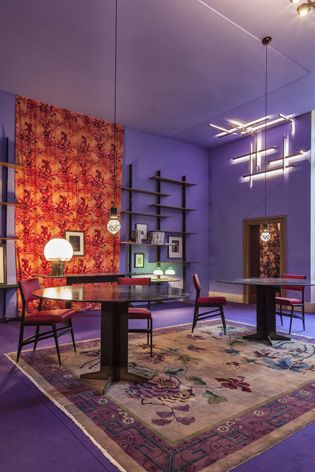 Room in purple tones with two glass-topped tables, red chairs, and a graphic light installation above the door