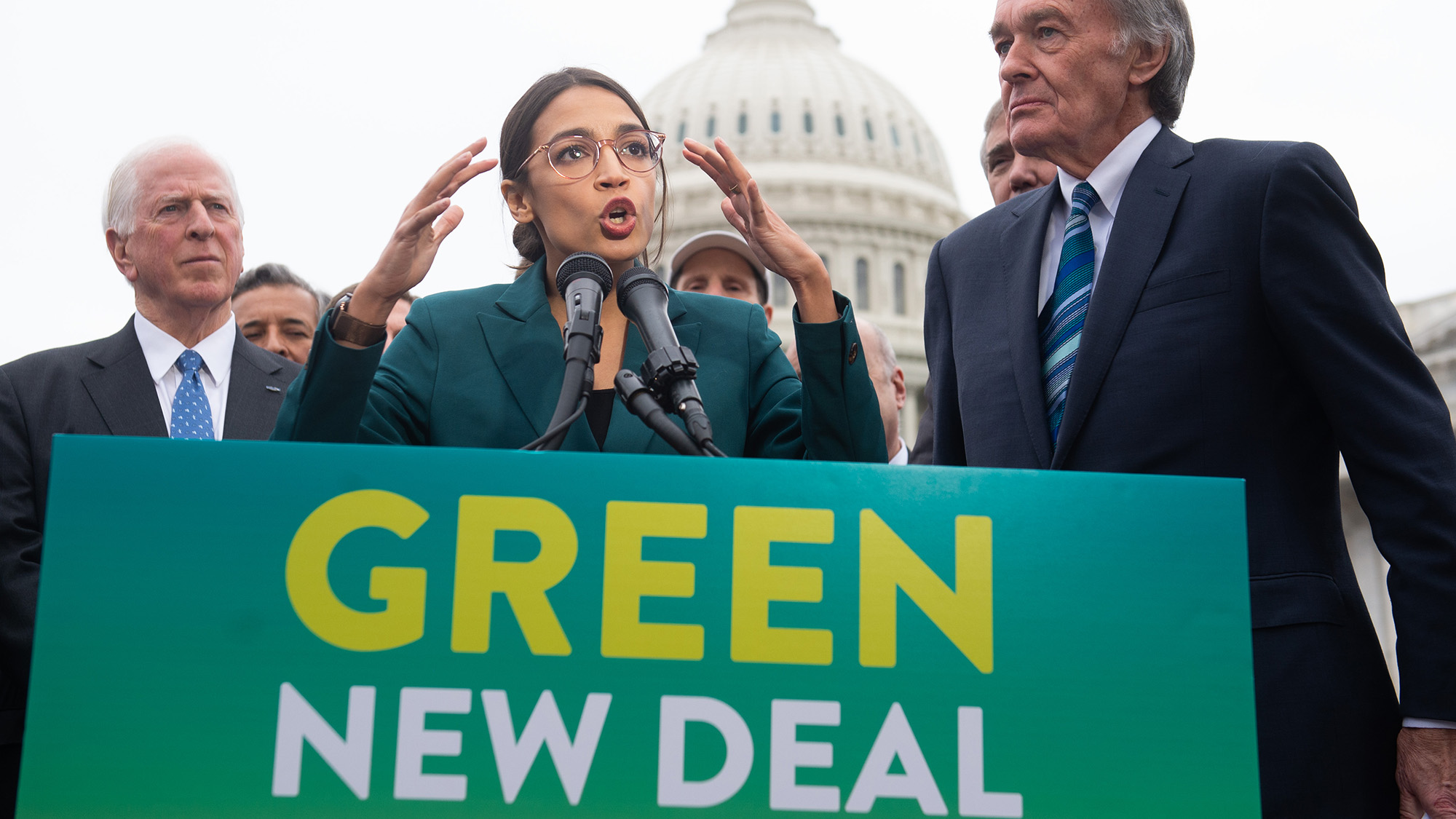 A woman with long dark hair and glasses speaks into a microphone behind a podium with a sign that reads "Green New Deal." 