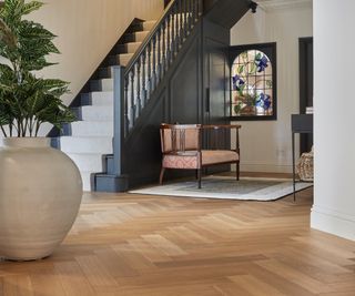 herringbone wooden flooring in large hallway with internal stained glass window, seating area, grey painted wooden stairs with cream runner and large cream plant pot and plant