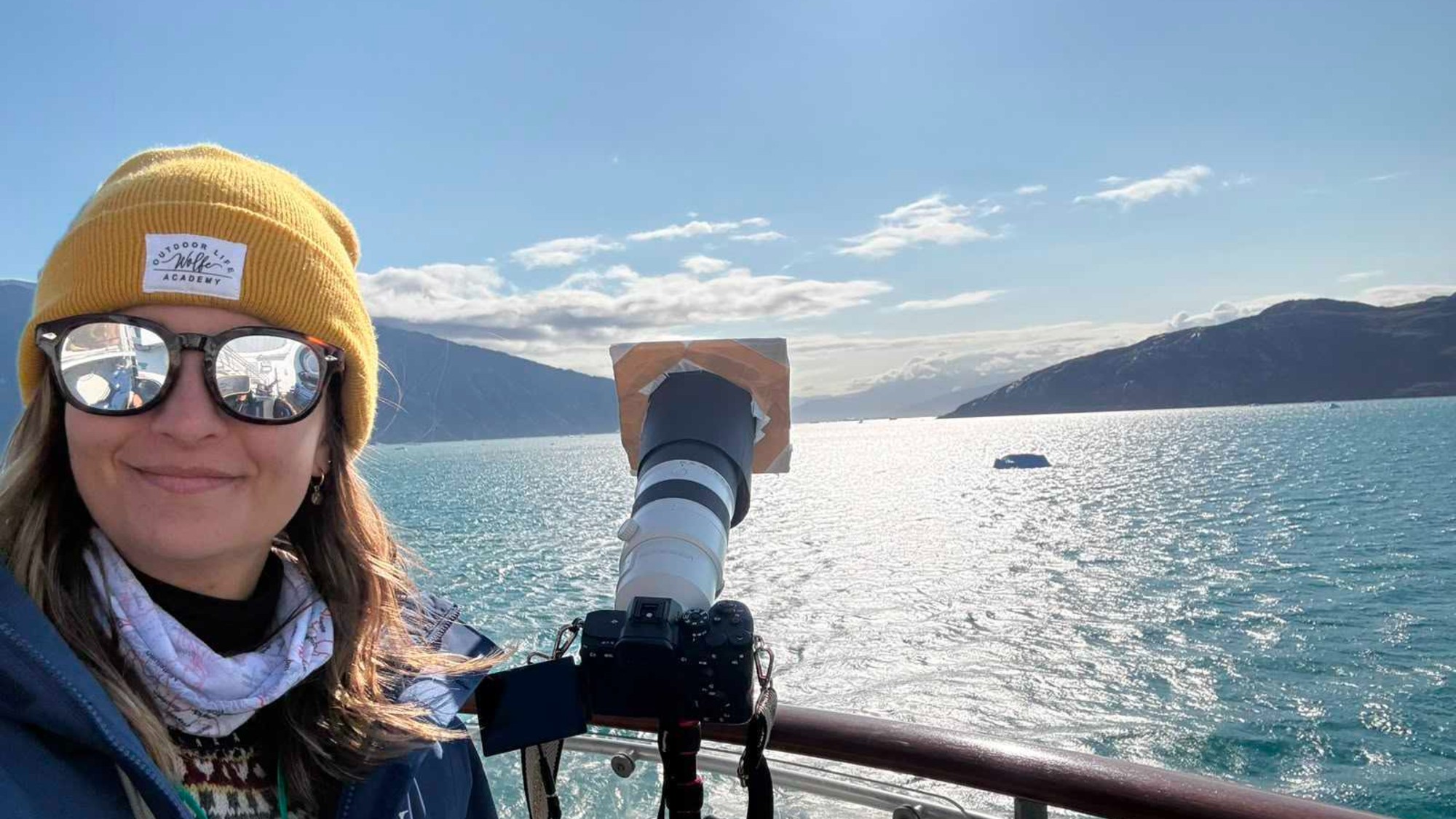 a woman in sunglasses and a winter hat poses beside a camera aboard a ship at sea