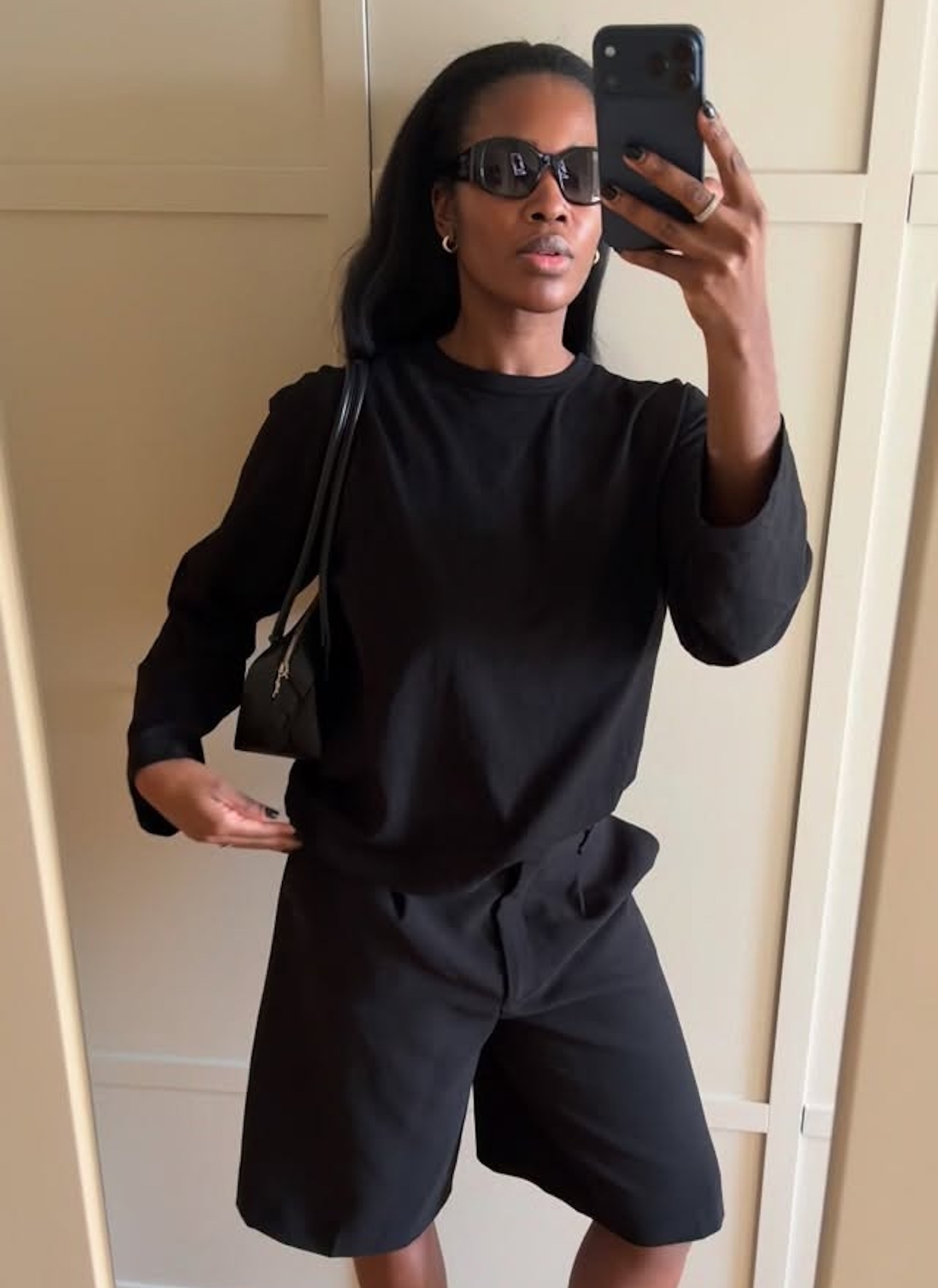 British style influencer Marilyn Nwawulor-Kazemaks poses for a mirror selfie in an all-black summer outfit featuring oversize sunglasses, a long-sleeve T-shirt, Bermuda shorts, and a shoulder bag.