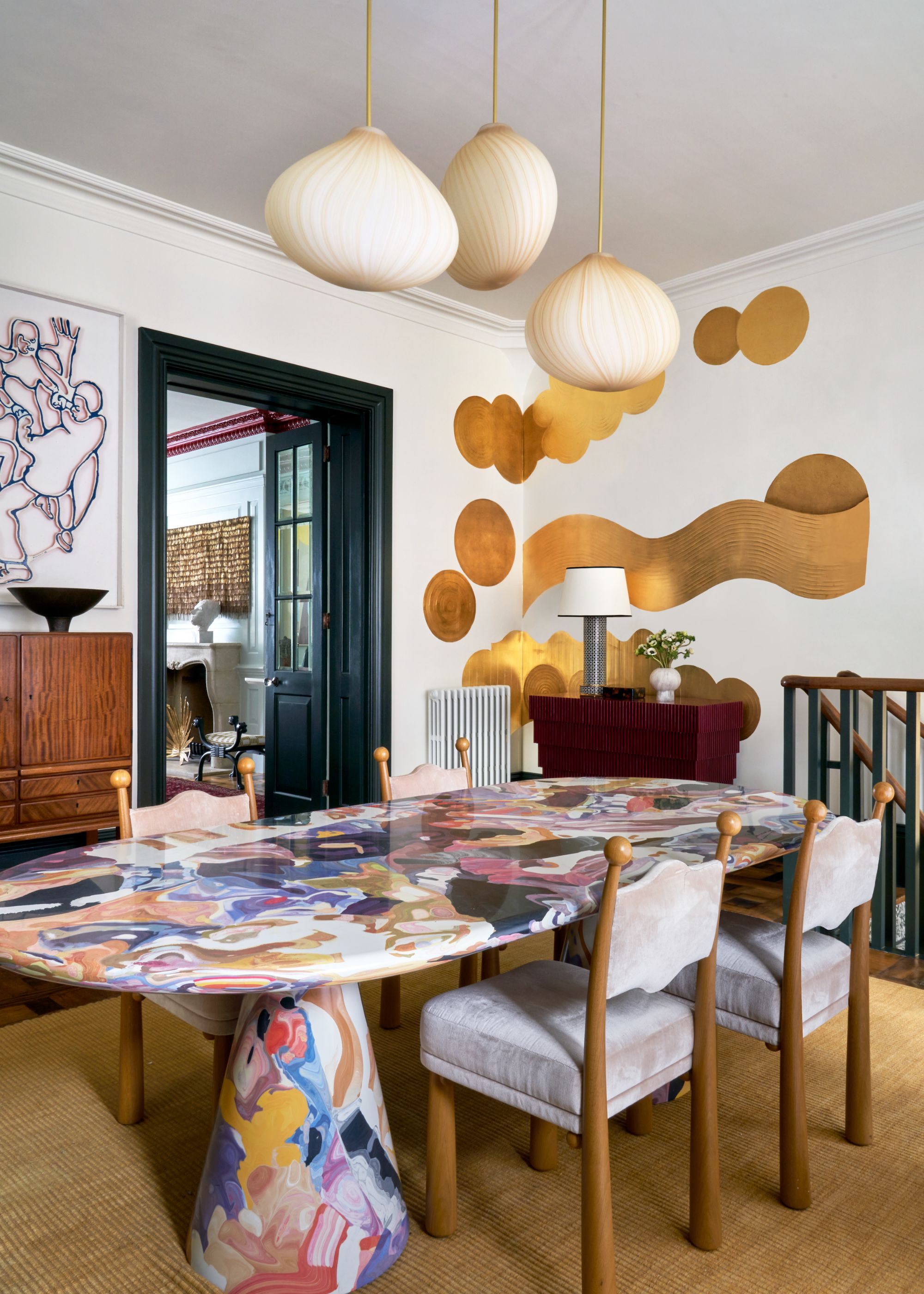 A playful dining room with white walls and an abstract gold wall mural, black trim, and a colorful patterned large dining table with wood and upholstered chairs. A jute rug and three sculptural pendant lights.