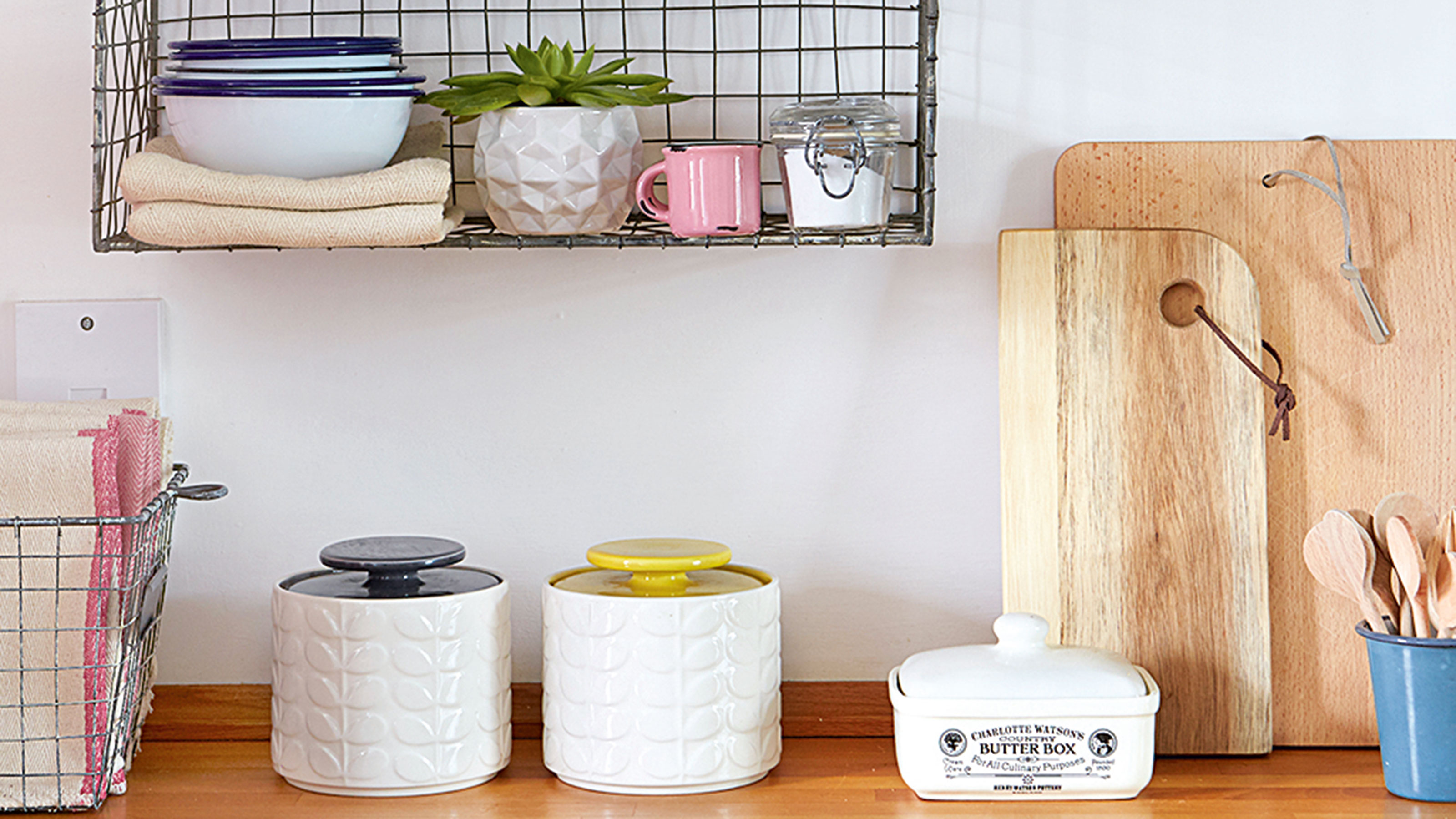 Wooden kitchen worktop with ceramic storage jars and a ceramic butter dish lined up against the wall next to two wooden chopping boards propped up