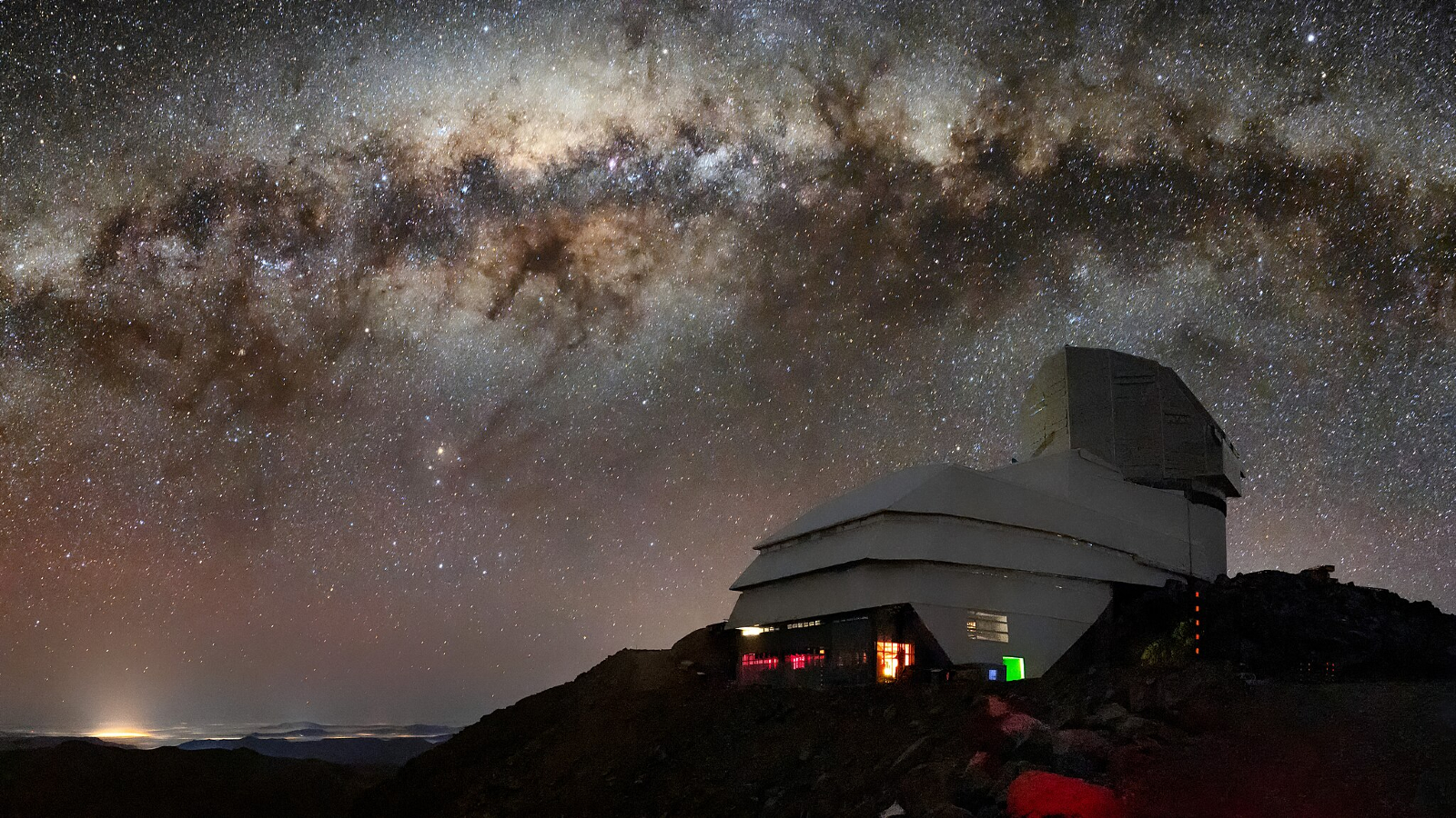 A glowing sky shows the Milky Way over a domed building on a cliffside