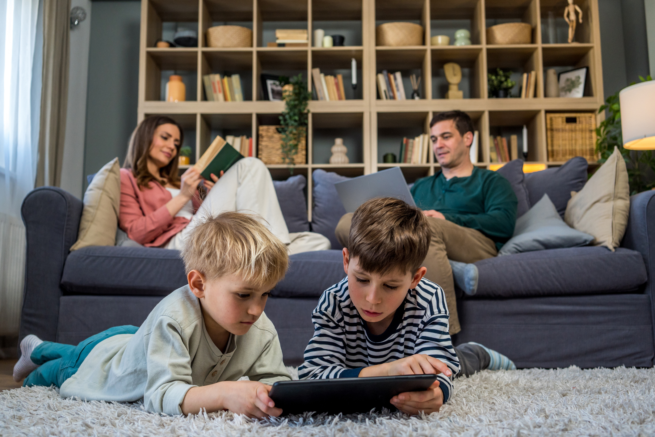 Two sons playing games on a tablet on the floor while their parents relax on the sofa with a laptop and a book, enjoying family time at home.