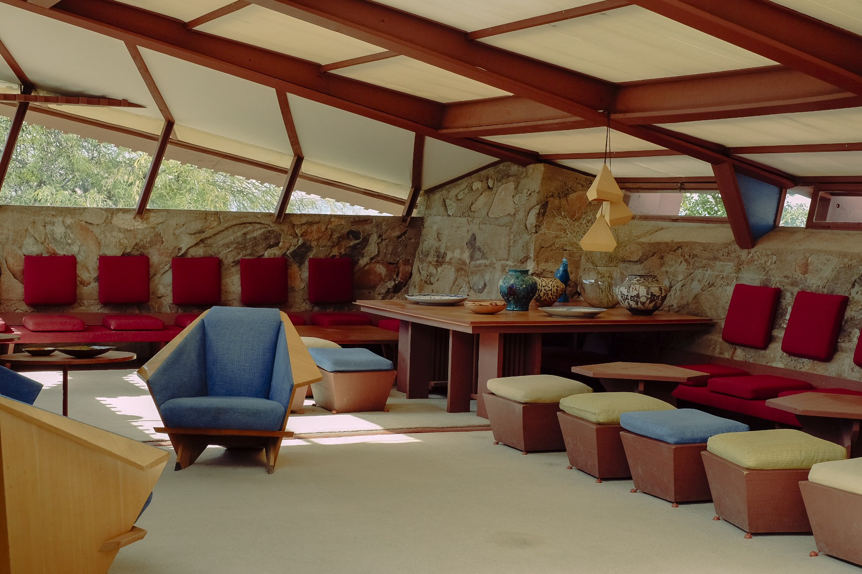 A modernist home immersed in the golden greenery of a desert features wood and textile ceilings in beige, carpeted floors in the same color, and geometrical furniture in tones of blue, acid green, bordeaux, and earthy browns.